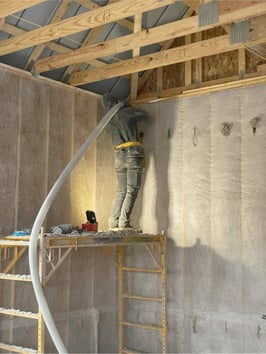 An insulation crew member standing on scaffolding while installing dense pack cellulose in the top of a wall cavity.