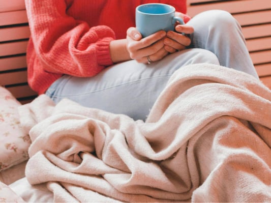 A woman in a sweater, under a blanket, holding a warm mug.