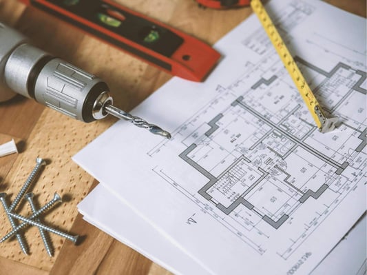 Blueprints for a floor plan on a wood table. The table also has screws, a drill, a level, and a measuring tape.