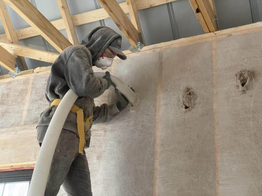 An insulation crew member installing dense pack cellulose behind netting into wood wall cavities in a metal garage building.