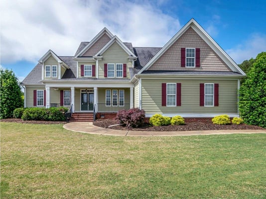 A two-story house with tan siding, landscaped bushes, trimmed green grass, and a concrete pathway to the front door.