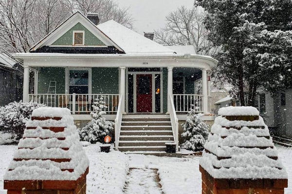 A house, wraparound porch, and front yard with several inches of snow.