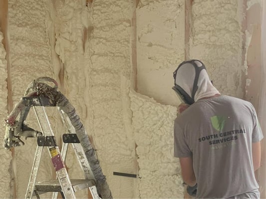 Trimming excess open cell spray foam from wall cavities in a new construction home. A ladder holds the inactive spray hose.