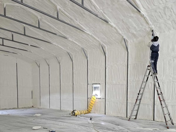 A spray foam contractor standing on a ladder to reach areas and scrape overspray.