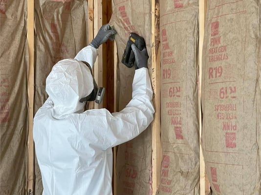 An insulation installer stapling R-19 fiberglass batts in place within wood cavities.