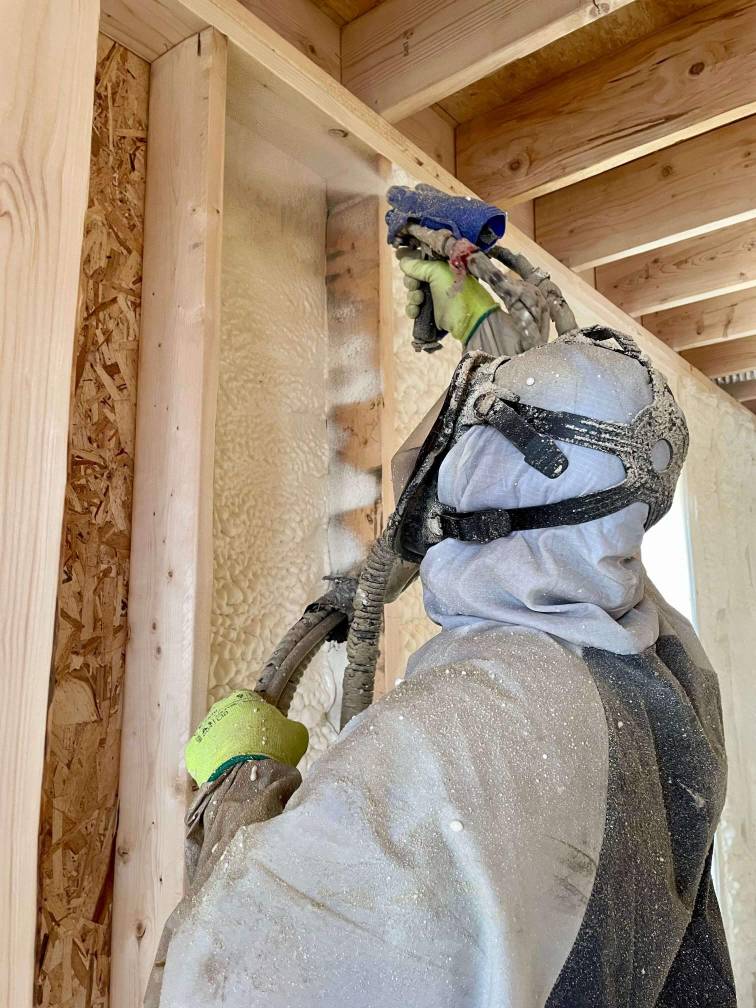 A spray foam contractor wearing a suit and ventilator, spraying open cell spray foam with spray gun applicator. Spray foam is being sprayed against a wood substrate wall cavity.